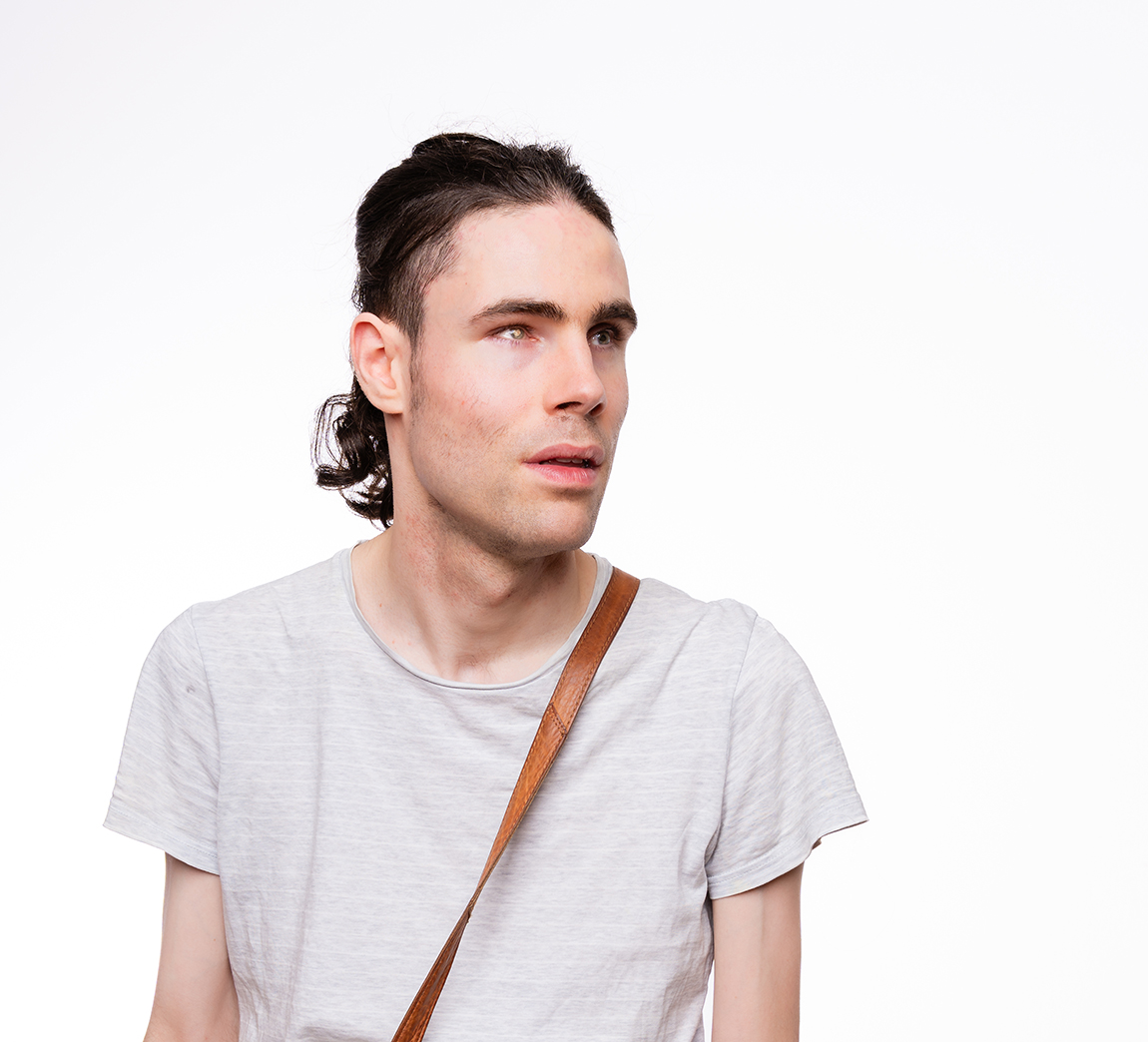 Vor weißem Hintergrund eine Portraitaufnahme von Dominic, der den Kopf zur Seite gewandt hat.