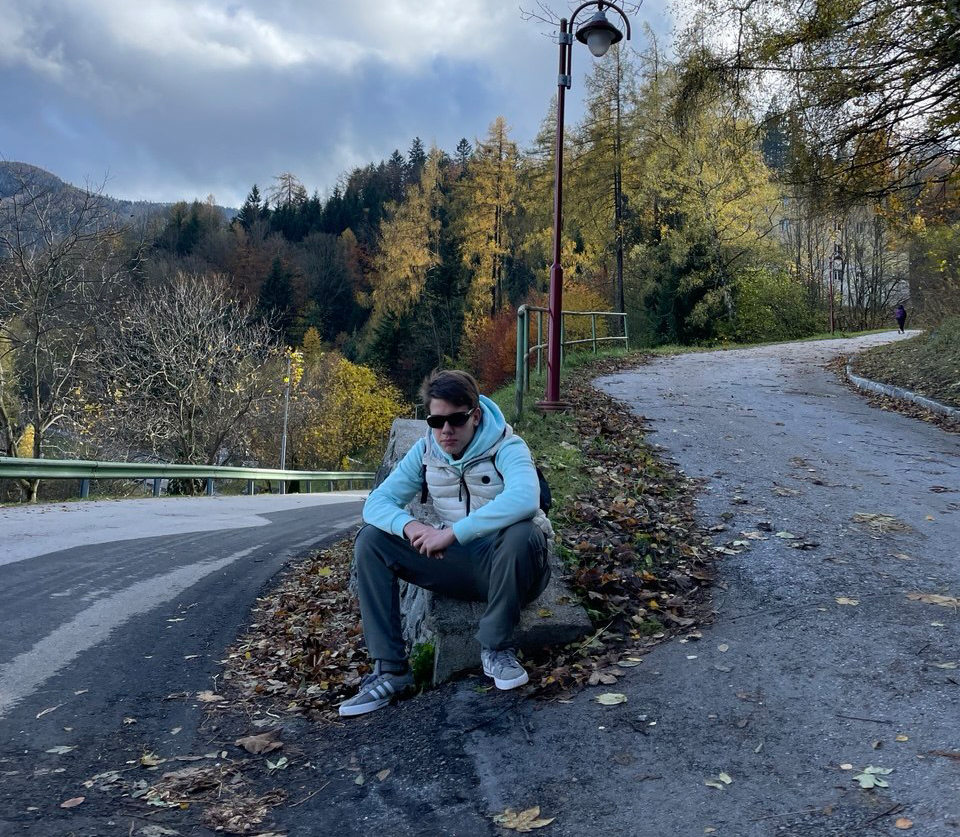 Ein junger Mann mit Sonnenbrille sitzt lässig auf einer Mauer zwischen zwei aufeinandertreffenden Bergstraßen, im Hintergrund Wald.