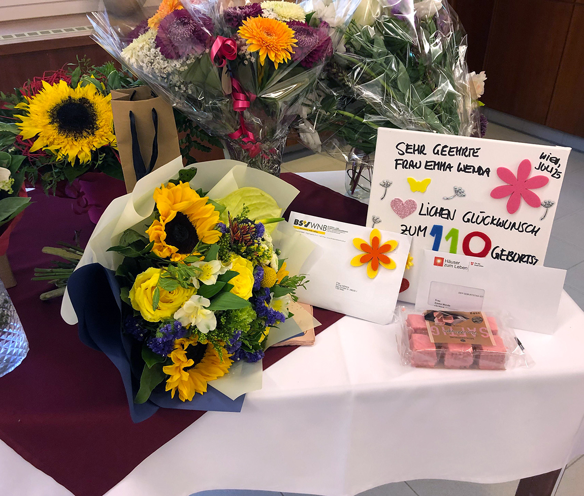 Auf einem Tisch mit weißer Tischdecke gelbe und bunte Blumensträuße, Punschkrapfen, eine Tafel mit bunt gestalteten Glückwünschen und ein Kuvert.