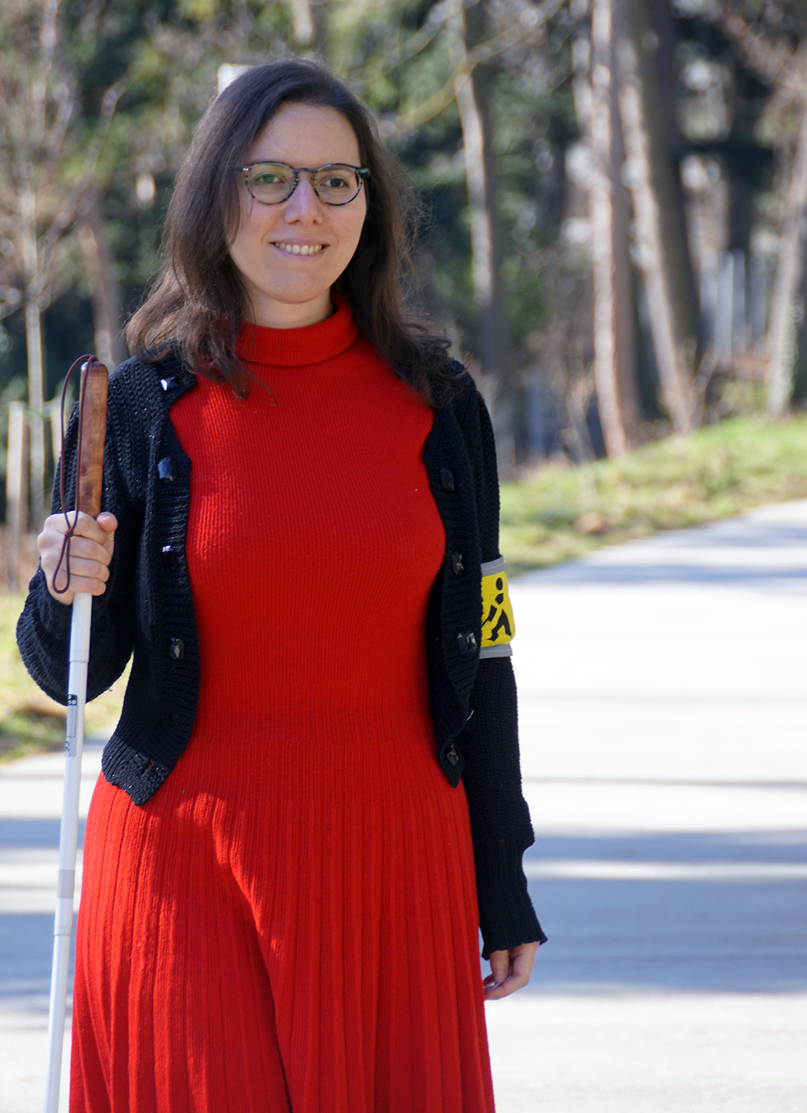 Eine lachende Frau mit schulterlangen dunklen Haaren und Brille steht auf einem breiten Gehweg in einem Park. Über einem roten Kleid trägt sie eine dunkle Strickweste, am linken Arm eine gelbe Schleife. In der rechten Hand ein Weißer Stock.