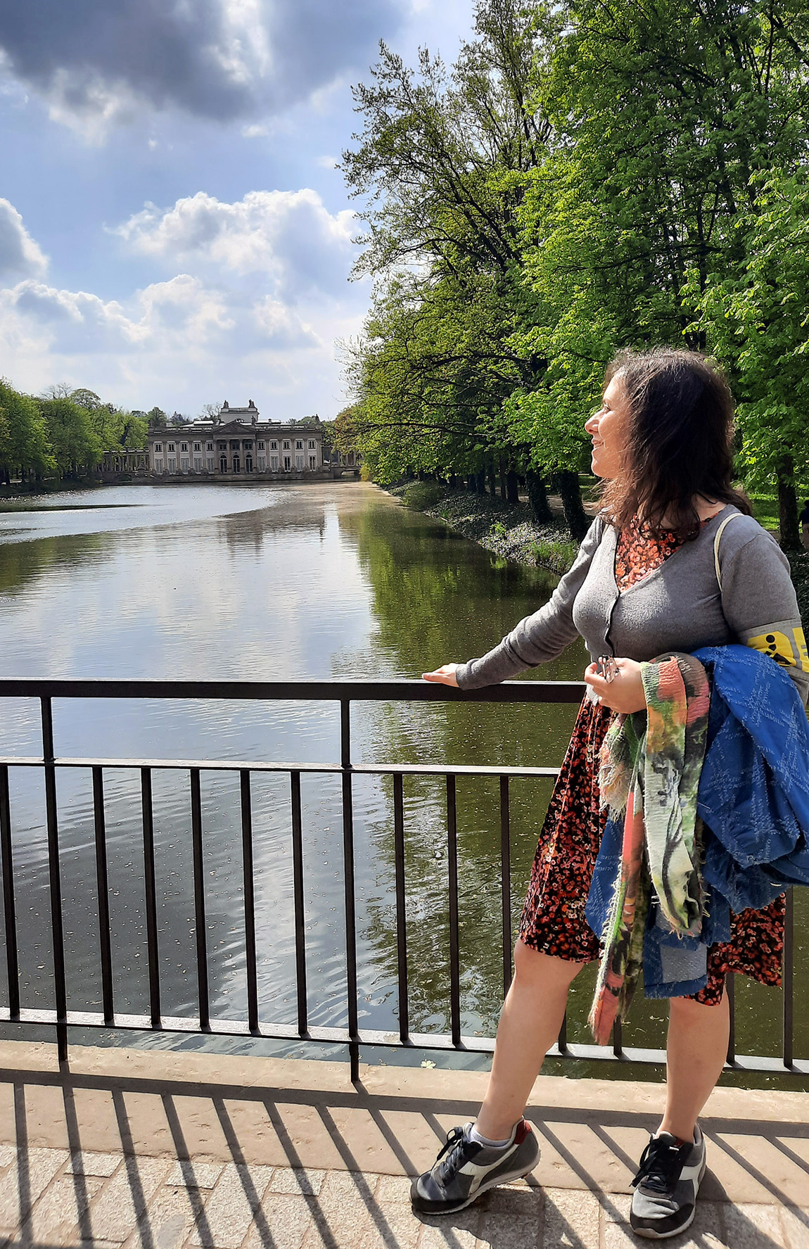 Eine Frau vor einem Geländer zu einem rechteckigen Teich hin. Am hinteren Teichufer steht ein breites Gebäude, an den seitlichen Ufern viele Bäume.