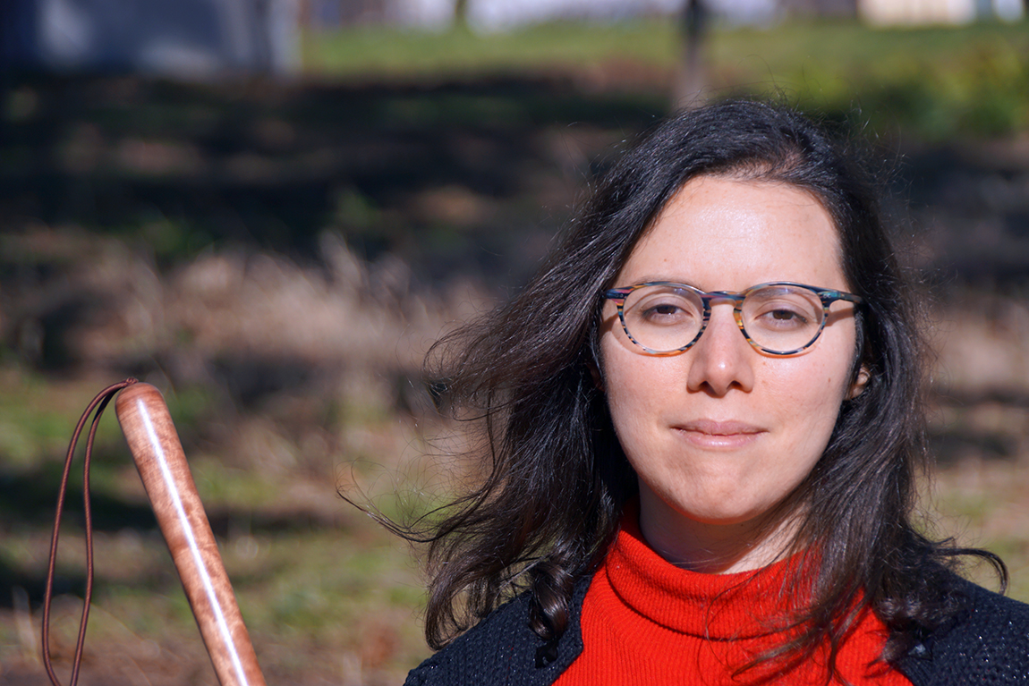 Portraitaufnahme einer Frau mit Brille und leicht im Wind wehenden Haaren. Links im Bild ist der Griff des Weißen Stockes zu sehen.