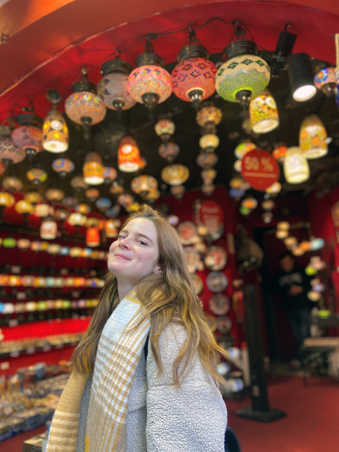 Eine lächelnde junge Frau mit langen Haaren vor einem Shop, an dessen Decke viele bunte Lampenschirme hängen.