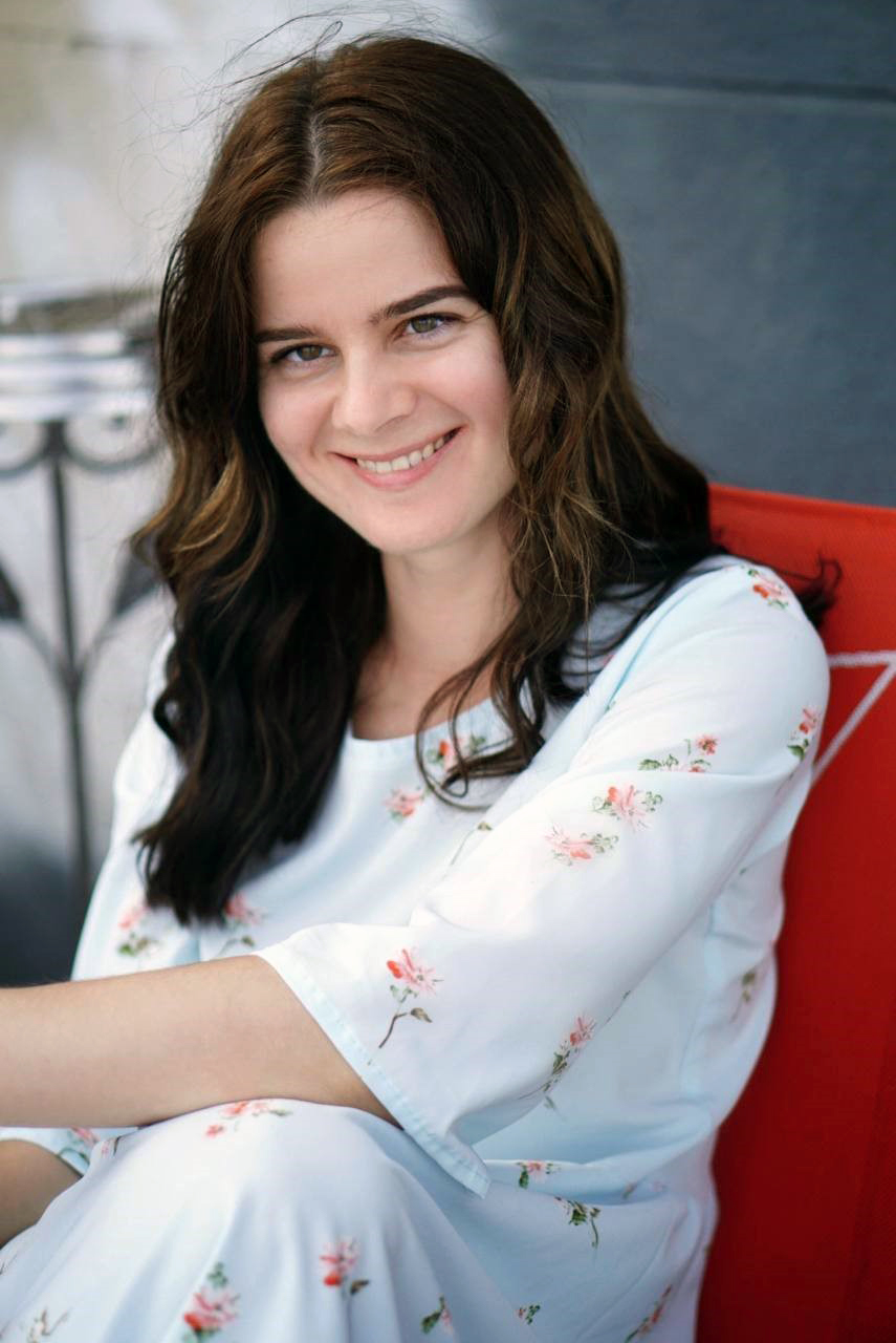 Eine in die Kamera lachende junge Frau mit langen hellbraunen Haaren, in einem weißen blumenverzierten Kleid.