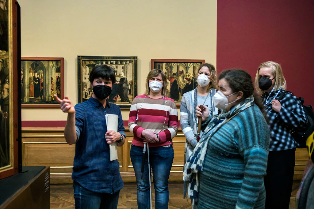 Fünf Frauen mit Mundschutz in einem Ausstellungssaal mit Gemälden an den Wänden.