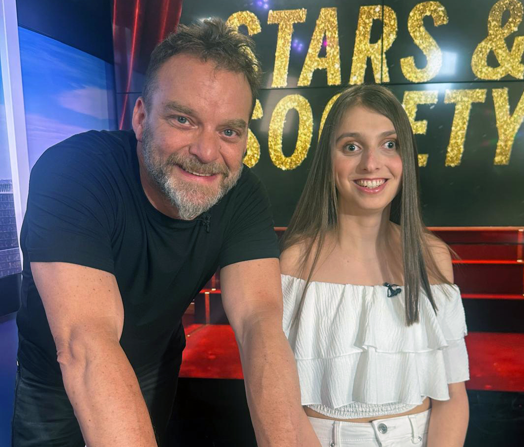 Ein Mann mit kurzen Haaren und Vollbart und eine junge Frau mit glatten langen Haaren lachen in die Kamera, im Hintergrund eine Tafel mit: STARS & SOCIETY